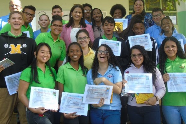 Group of smiling students at Filomena Mavo College proudly holding certificates after an award ceremony, surrounded by teachers and staff.