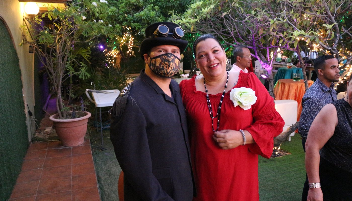 Teacher (Man) in a steampunk costume and the Dean of Filomena Mavo (woman) in a red dress with a flower accessory posing at a Filomena Mavo College graduation evening party.