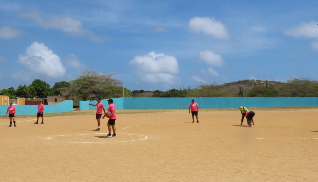 Filomena Mavo College students and teachers playing a kickball game on a dirt field under a blue sky, wearing bright pink uniforms.