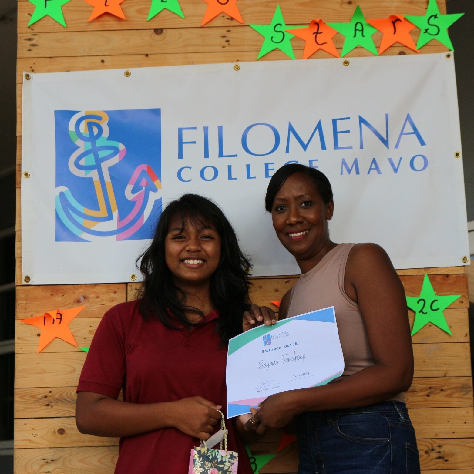 A smiling student in a red polo shirt receiving a certificate for academic excellence from a teacher at Filomena College MAVO. They stand in front of a banner with the school’s logo, decorated with colorful stars.