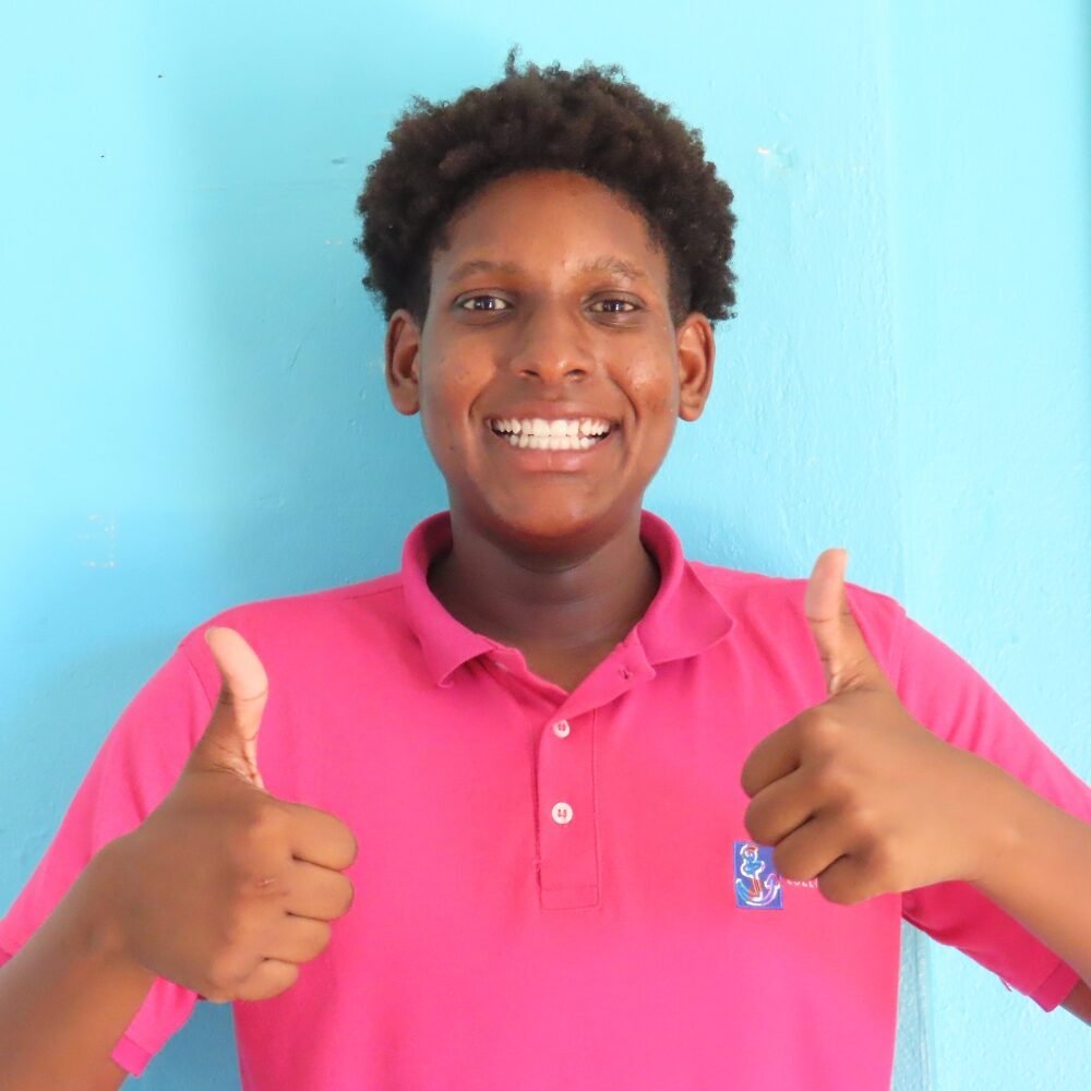 A smiling student wearing a pink polo shirt with the Filomena College MAVO logo, standing against a bright blue wall, giving two thumbs up.